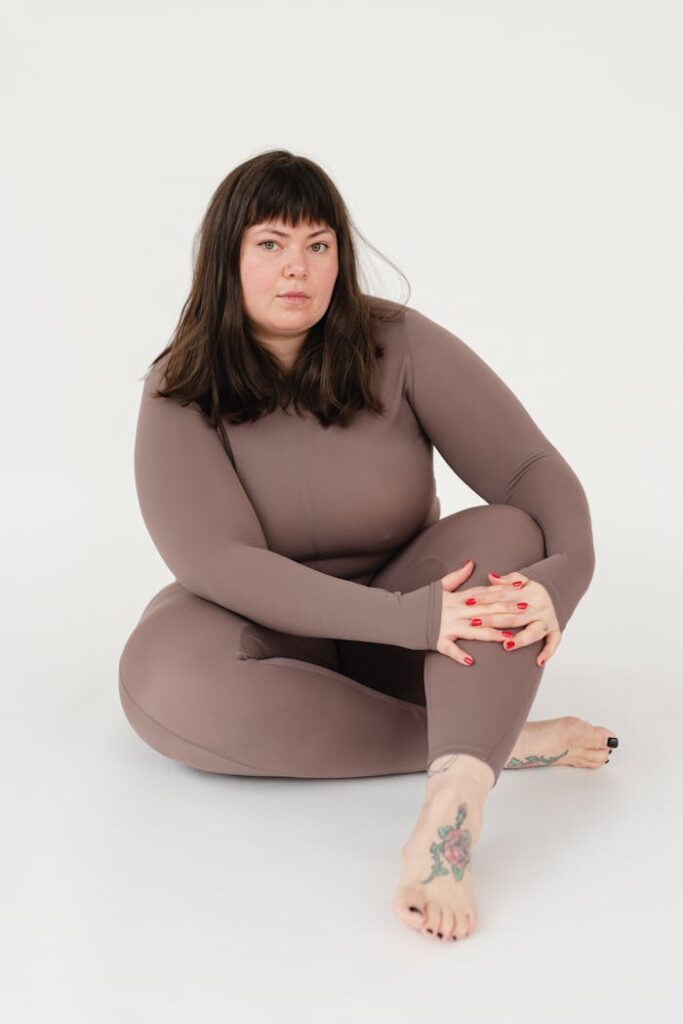 Full body of serious overweight female in sportswear embracing knee while sitting with legs crossed against white background and looking at camera